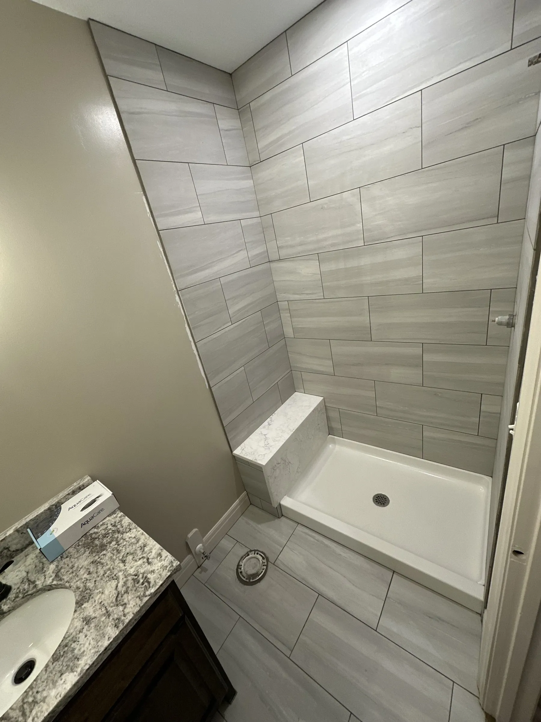 A shower tiled in gray wood-look porcelain with a bench and a white base, beside a dark vanity