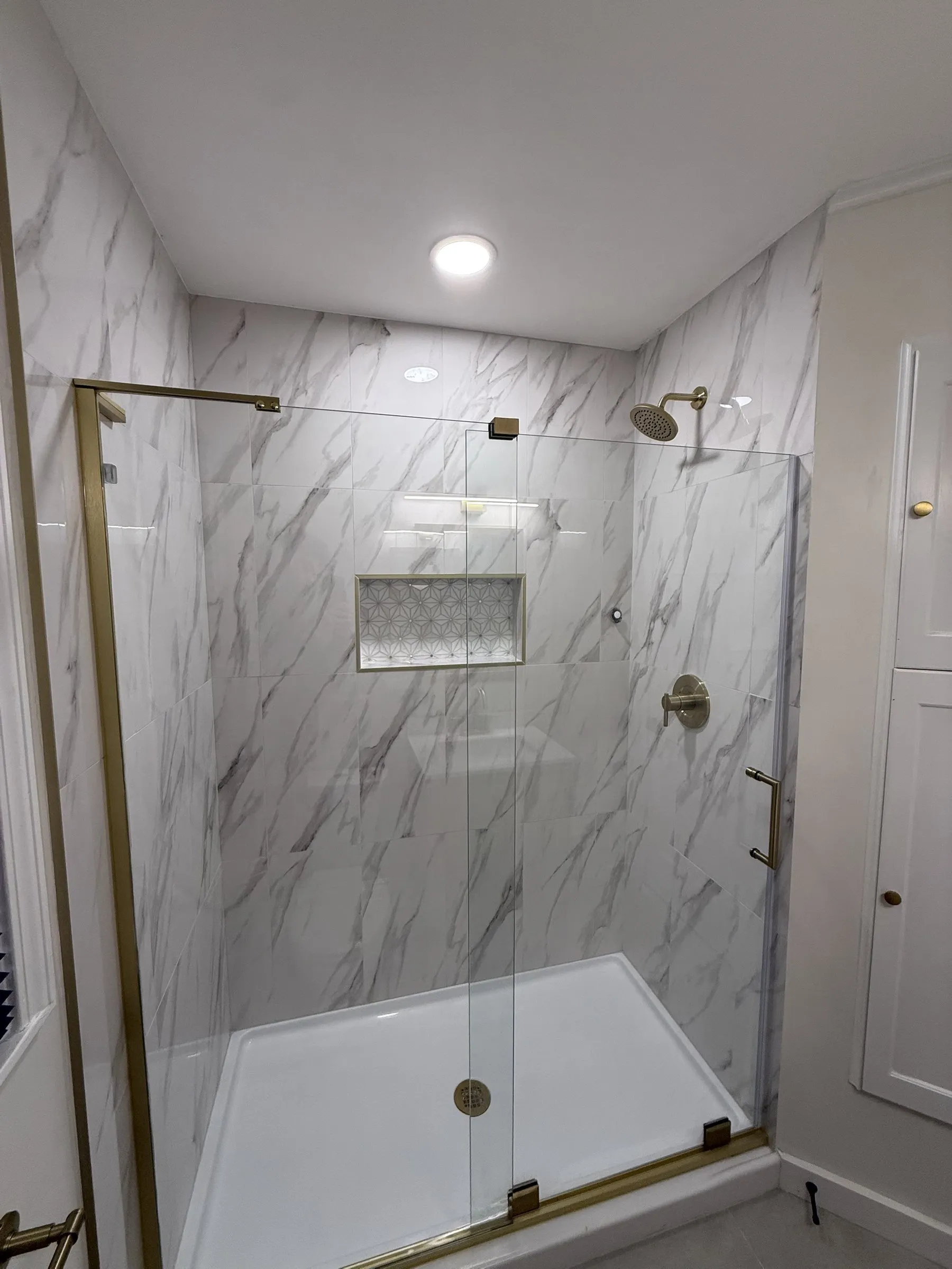 A walk-in shower tiled in marble-look porcelain with a patterned niche, a glass door and brushed brass fixtures