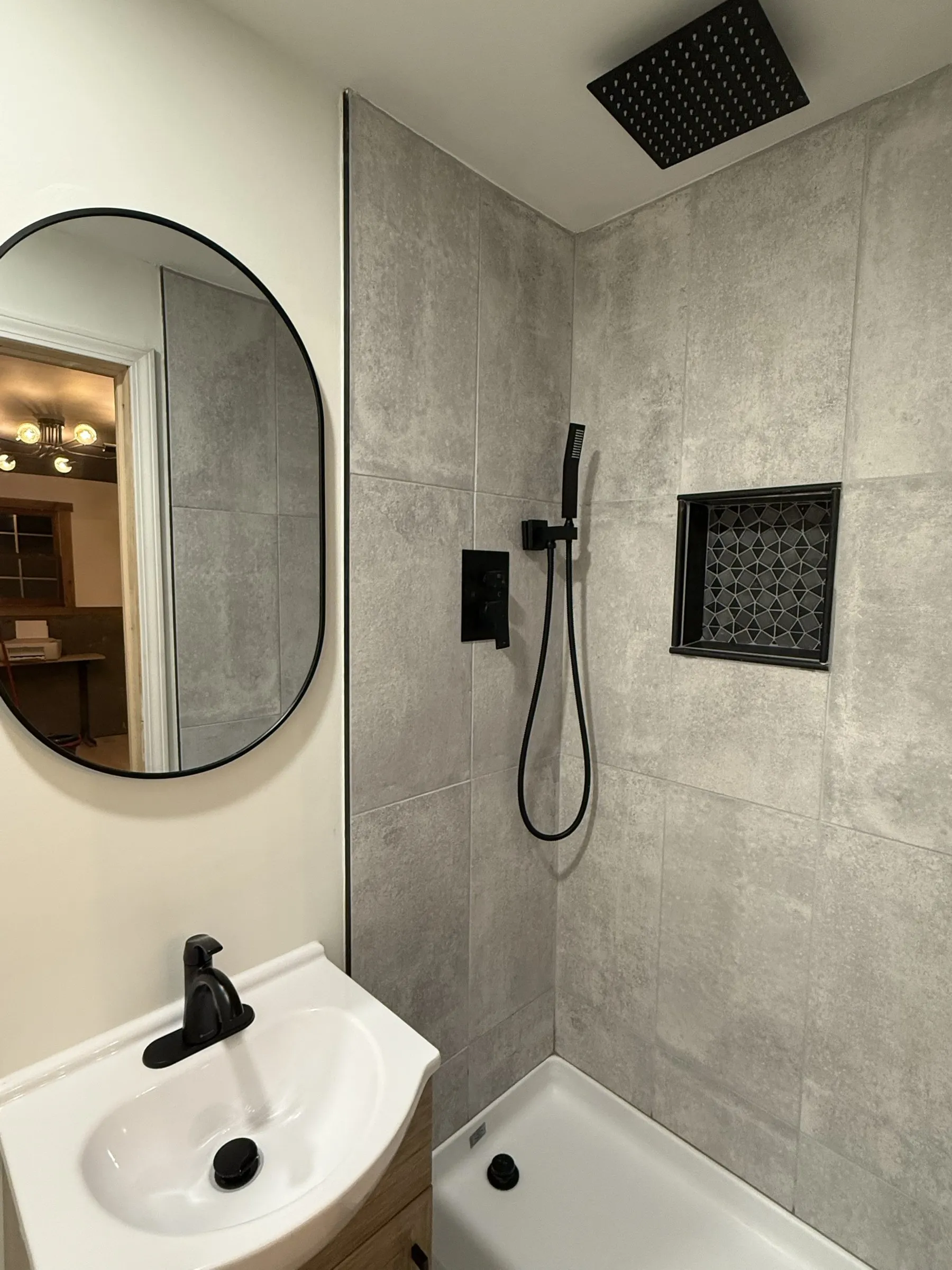 A small bathroom with concrete-look tile, a matte black rain head, a round mirror and a wood vanity