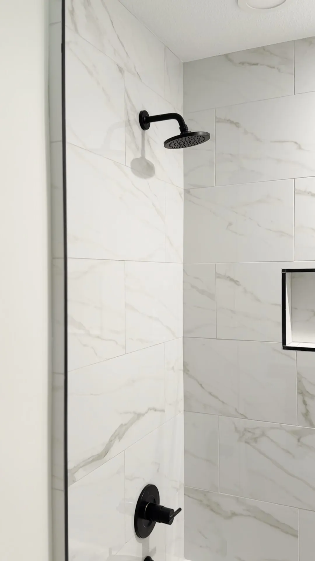 A shower tiled in marble-look porcelain with a matte black shower head and a black-framed niche
